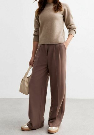 Kvinde iført beige sweater, brune bukser med vide ben, beige sneakers, holder en lille beige håndtaske med den ene hånd i lommen.