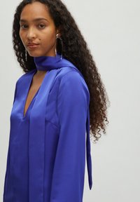 Blauwe satijnen blouse met een diepe V-hals, lange mouwen en een strikdetail bij de hals. Gladde textuur, losse pasvorm en minimale stiksels.