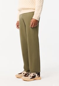 Olijfgroene broeken met een rechte pasvorm, gemaakt van gladdig materiaal; gecombineerd met beige en bruine sportschoenen met accenten en meshpanelen.