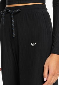 Black ribbed sweatpants with a drawstring waist, featuring blue-striped accents and a small heart logo embroidered on the leg.