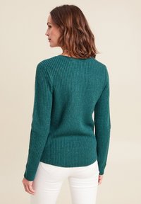 Maglione lavorato a maglia color turchese con texture a costine, scollatura rotonda e maniche lunghe. Presenta un orlo leggermente curvato ed è abbinato a pantaloni bianchi.