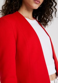 Rode blazer met een gladde textuur, open voorkant en geen zichtbare sluitingen. Draag het over een witte top, wat een casual uitstraling geeft.