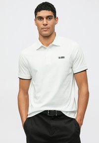 Férfi fehér rövid ujjú pólót visel, kicsi fekete logóval a bal mellkasán, fekete nadrágban, kezében a zsebébe dugva áll egy sima háttér előtt.