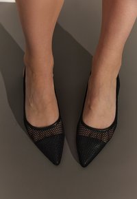 Baletăne negre cu vârful ascuțit, având un superior din plasă cu un model texturat și accente din piele netedă. Un design elegant și aerodinamic.