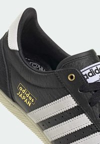 Black leather sneaker with white suede three stripes, gold "adidas JAPAN" logo, textured upper, and patterned rubber sole.