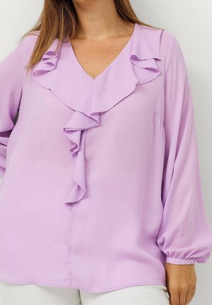 Kvinde iført en lyse lilla bluse med flæset V-halsudskæring og lange ærmer med elastiske manchetter.