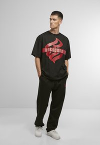 Juodas trumpomis rankovėmis marškinėliai su raudonu flame logotipu ir "ROCWEAR" tekstu, derinami su juodomis sportinėmis kelnėmis ir baltais sportbačiais. Kasdienis apranga.