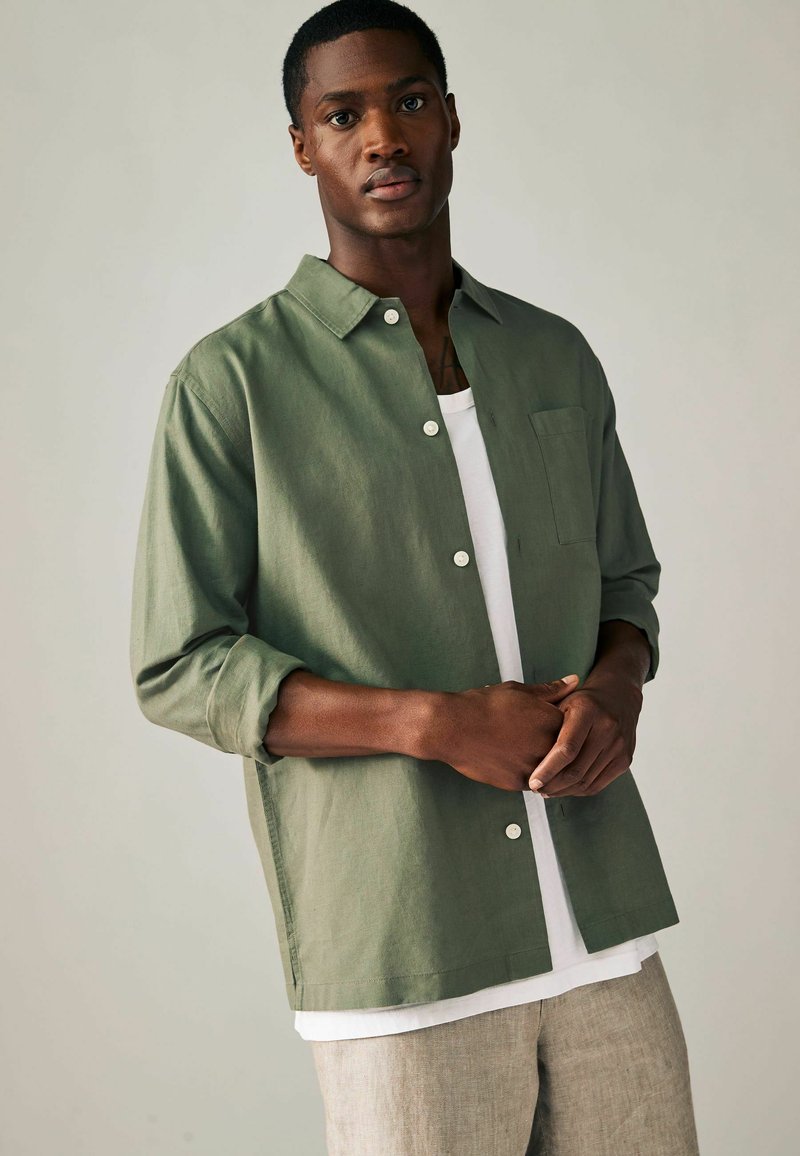 Olivno zelena, srajca z dolgimi rokavi z gumbi, z žepom na prsih, belimi gumbi in sproščenim krojem, oblečena preko belega topa brez rokavov.