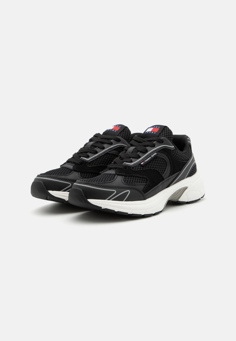 Baskets de sport noires en matériaux mesh et suède, avec une semelle rembourrée, des accents réfléchissants et un logo bien visible sur la languette.