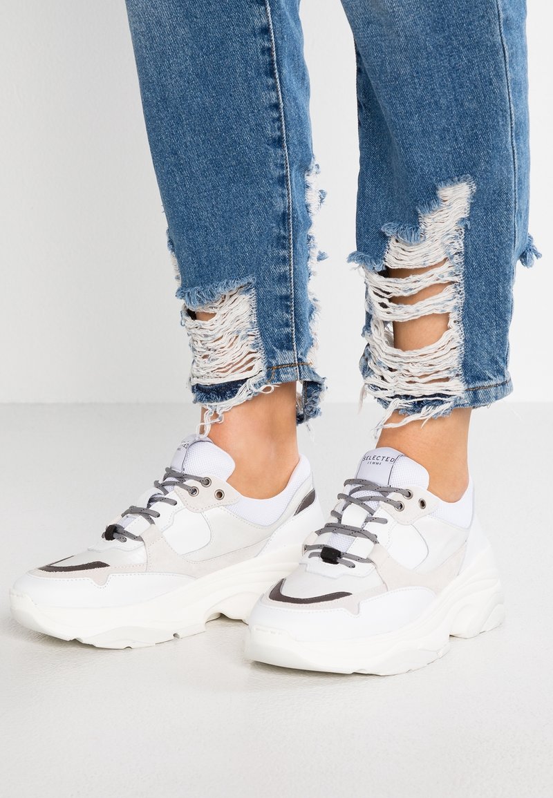 Baskets blanches et gris clair avec un design superposé, des matériaux texturés et des accents noirs. Portées avec un jean bleu effiloché avec des déchirures au niveau des chevilles.