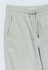 Pantalon beige clair avec taille élastique et cordon, avec une grande poche plaquée à l'avant et une poche fendue latérale d'un côté.