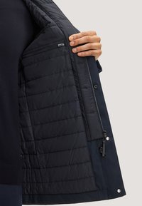 Marine geïsoleerde jasinterieur met gevoerde voering, ritszak en drukknoopdetails. Zachte textuur, rechte snit, wat bijdraagt aan de warmte.