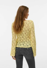 Top in pizzo giallo con maniche lunghe e motivo floreale, caratterizzato da un collo rotondo e una texture semitrasparente. Indossato con jeans grigi.
