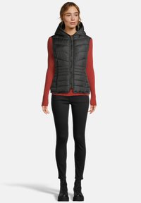 Vrouw in een zwarte gewatteerde vest met capuchon over een rood geribbeld longsleeve shirt, zwarte skinny broek en zwarte veterschoenen, rechtopstaand en naar voren kijkend.