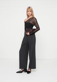 Top nero trasparente a maniche lunghe, pantaloni grigi a vita alta e a gamba larga, e scarpe nere con tacco a blocco. Clutch bordeaux con dettagli metallici.