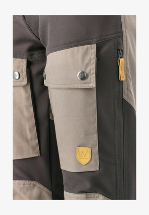 Cargo bukser i mørkegrå og beige stof, med flere lommer, sølv knaplukninger og et gult læderlogo.