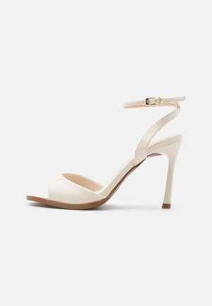 Beige sandaal met hoge hak, dunne enkelband met gouden gesp, open teen en slanke naaldhak op een witte achtergrond.