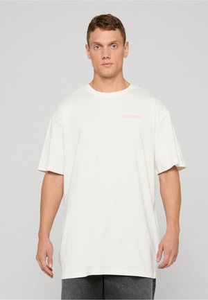 Junger Mann mit kurzen braunen Haaren, der ein übergroßes einfarbiges weißes T-Shirt und graue Hosen trägt, vor einem einfarbigen Hintergrund stehend.