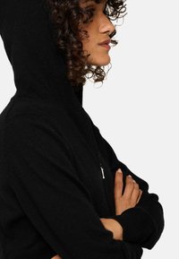 Haut de sweat à capuche zippé noir avec un col haut et un petit tirette. Il présente une texture douce et des manches longues, s'ajustant étroitement au corps.