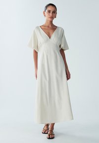 Maxikleid aus leicht beigem Stoff mit V-Ausschnitt, kurzen weiten Ärmeln und tailliertem Oberteil, das eine glatte Textur und ein minimales Design aufweist.