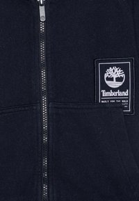 Jūras spēku zips-vilkšanas kapuce ar mīkstu tekstūru, ar priekšējo kabatu un balto Timberland logotipa uzšuvumu kreisajā krūtī. Zips ir metalizēts.