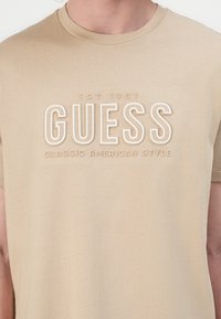 Beige bomull t-skjorte med hevet hvit tekst "GUESS" og "CLASSIC AMERICAN STYLE" med "EST 1981" over, korte ermer og rund halsåpning.