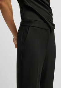 Zwart op maat gemaakte broek van een gladde, textuur stof, met een slim fit ontwerp en zijzakken. Details omvatten een platte voorkant en subtiele plooien.