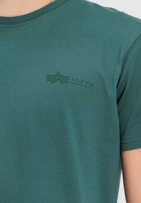 Krátký rukáv zelené bavlněné tričko s kulatým výstřihem, s jemným potiskem loga v tmavší zelené barvě na levé straně hrudi.