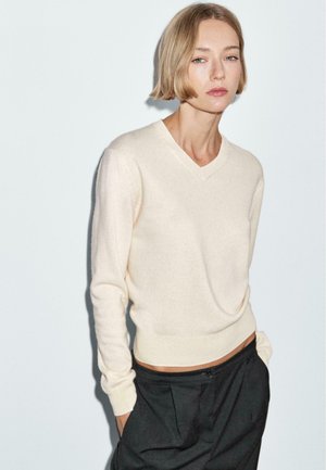 Maglione crema a V in tessuto morbido con maniche lunghe, silhouette aderente e orlo a coste. Abbinato a pantaloni neri su misura.