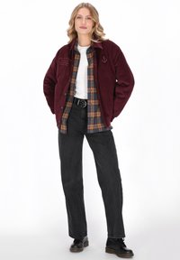 Giacca in corduroy bordeaux, abbottonata, abbinata a una camicia flanella a quadri nei toni del marrone e blu navy. Jeans neri a gamba dritta e stivali neri.