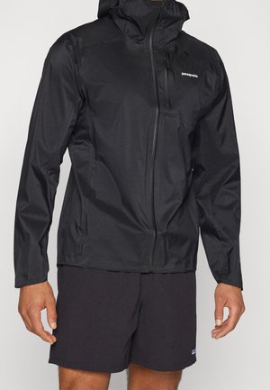 Sort Patagonia hættejakke med lynlåslomme på brystet foran, fuld lynlåslukning, båret med sorte shorts på en stående person.