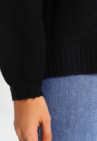 Close-up van een hand met een zwarte gebreide trui mouw, rustend op blauwe denim jeans.