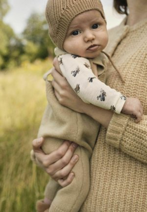Beige'ist kootud müts ja kehakate seljas beebi, keda hoiab beige kampsunis inimene, väljas, uduste muru ja puude taustal.