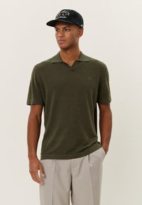 Dark green knit polo shirt with a V-neck and short sleeves, featuring a subtle logo on the left chest. Worn with light striped pants.