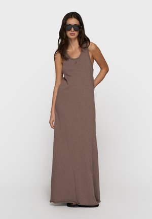 Kobieta w brązowej sukience maxi bez rękawów i dużych czarnych okularach przeciwsłonecznych, stojąca na tle jednolitego jasnego tła.