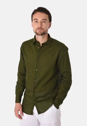 Uomo con capelli corti e scuri e barba, indossa una camicia verde scuro con bottoni e pantaloni bianchi, in piedi con una mano in tasca.