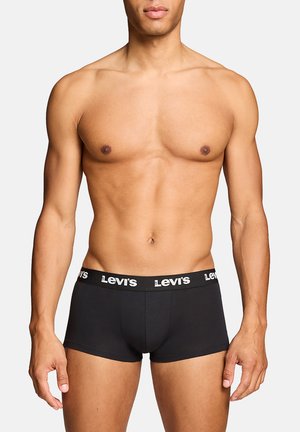 Man with muscular build wearing black Levi's boxer briefs, standing against a plain light background with arms relaxed by sides.