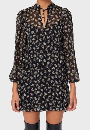 Mujer que lleva un vestido mini negro con estampado floral, mangas largas transparentes y escote con lazo en forma de ojo de cerradura, combinado con botas negras hasta la rodilla.