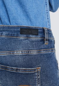 Jeans in denim di blu medio con un'etichetta nera del marchio sulla tasca posteriore. Presentano cuciture arancioni e un design a gamba dritta.