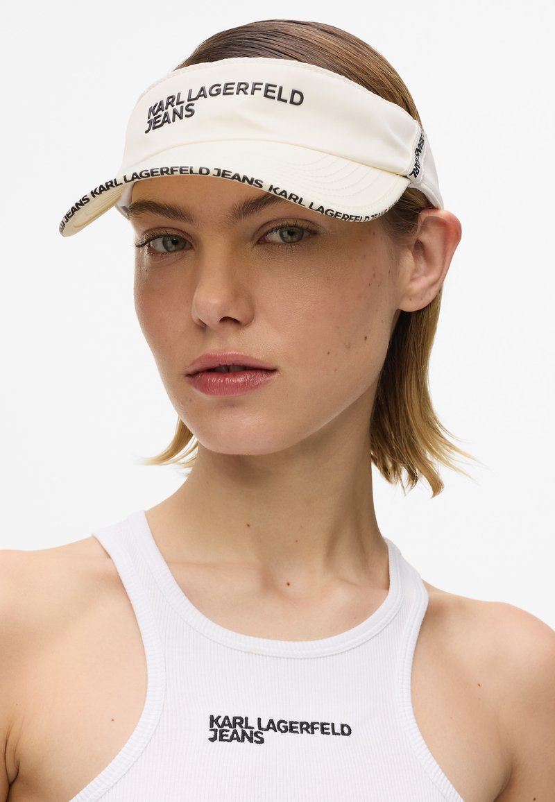 Casquette visière blanche avec le logo noir « KARL LAGERFELD JEANS », dotée d'une visière courbée et d'une texture de tissu lisse, portée avec un débardeur blanc.