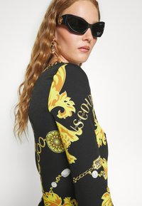 Top negro de manga larga con atrevidos patrones barrocos amarillos, detalles en dorado y un diseño ajustado. Los accesorios incluyen gafas de sol negras y pendientes.