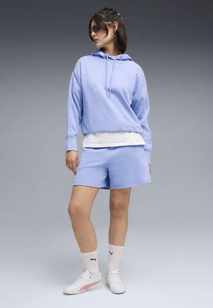 Mujer con sudadera con capucha azul claro y pantalones cortos a juego, camiseta blanca en capas, calcetines blancos de Puma, zapatillas blancas y gafas de sol, de pie frente a un fondo gris.