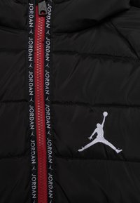 Sort polstret jakke med rød lynlås, sort lynlås tape med gentagen hvid "JORDAN" tekst og hvid Jumpman logo broderi.