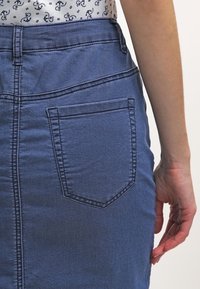 Vista trasera de una persona que lleva una falda de mezclilla azul con un bolsillo trasero y una camisa blanca con un patrón de anclas marinas en color navy, con el brazo derecho relajado a un lado.