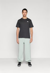 Σκούρο γκρι μπλουζάκι με λευκό λογότυπο, συνοδευόμενο από ανοιχτό μέντα πράσινο παντελόνι jogging. Μαύρα αθλητικά παπούτσια φορεμένα σε απλό φόντο.