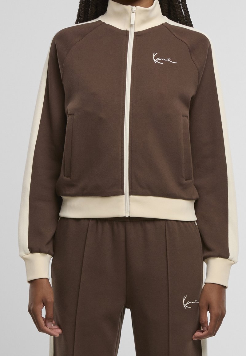 Personne portant une veste de survêtement marron et crème zippée et un pantalon assorti avec un logo blanc sur la poitrine et la cuisse.