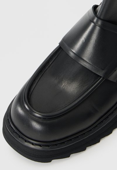 Mocassin en cuir noir avec une finition lisse, embout rond et une sangle sur le cou-de-pied. Semelle en caoutchouc épaisse avec des coutures décoratives.