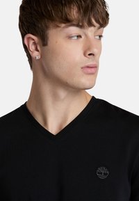 T-shirt nera con scollatura a V realizzato in cotone con una texture morbida. Presenta un discreto logo grigio di un albero sul petto, design aderente.
