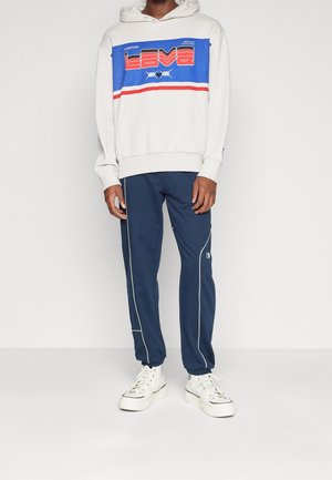 Biała bluza z kapturem z niebieskim, czerwonym i białym graficznym wzorem, zestawiona z granatowymi spodniami dresowymi z białymi lamówkami i logo. Białe sneakersy.