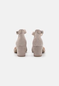 Une paire de sandales à talons blocs en daim beige avec des brides de cheville et des boucles argentées, présentées de dos sur un fond blanc.